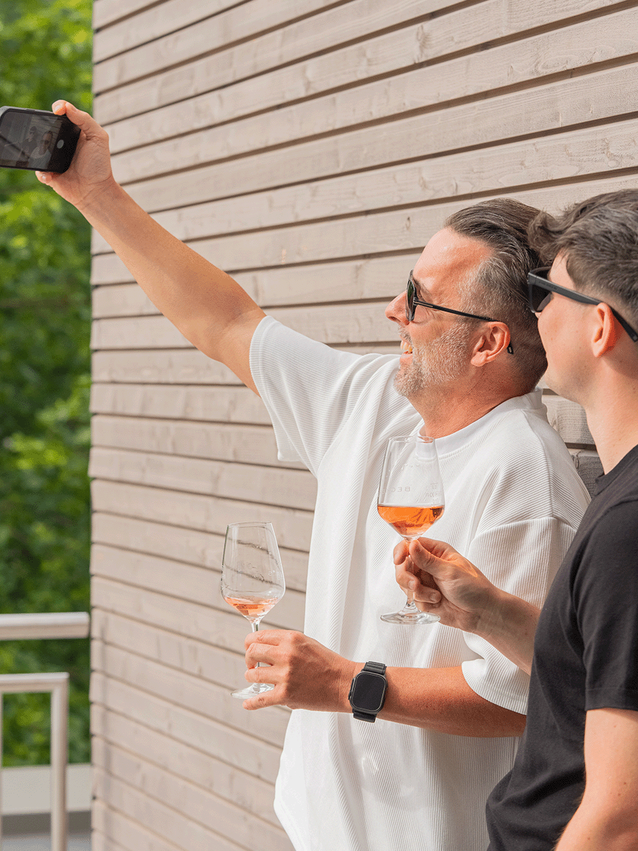 Zwei Männer mit Sonnenbrillen stehen nebeneinander und halten Weingläser, während einer ein Smartphone für ein Selfie hochhält