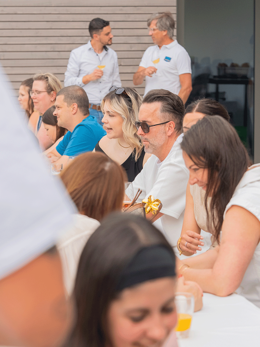 Gruppe von Menschen sitzt an einem langen Tisch und unterhält sich, im Hintergrund zwei Männer stehend im Gespräch